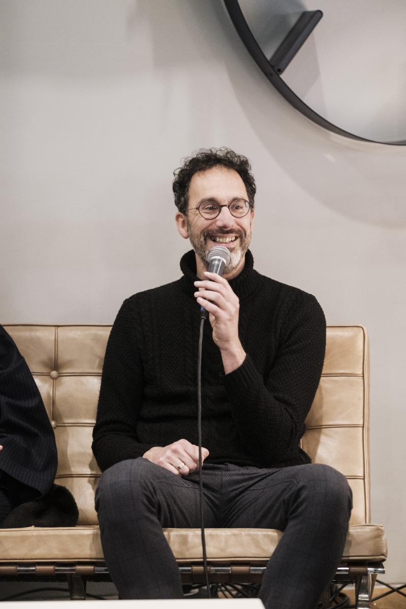 Leerkracht Piet Creten, een man in het zwart gekleed zittend op een sofa, microfoon in de hand