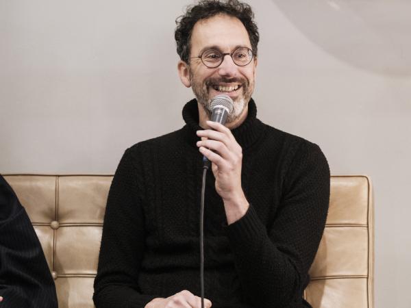 Leerkracht Piet Creten, een man in het zwart gekleed zittend op een sofa, microfoon in de hand